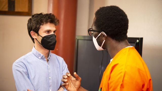 Omar Alshogre (left) wears a mask and a blue button down and talks with a scholar in GA&#039;s Prison Scholars Program who wears an orange jumpsuit and a mask.